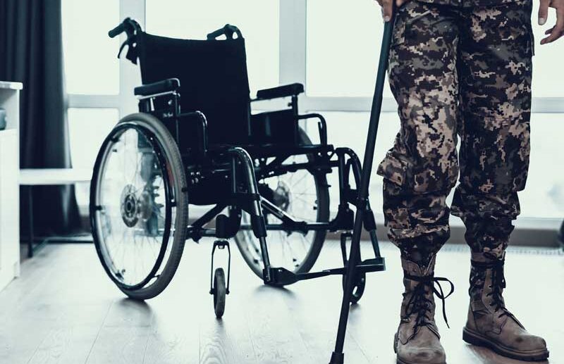 the bottom half of a picture of a person in military fatigues holding a cane to stand and there is a wheelchair behind him