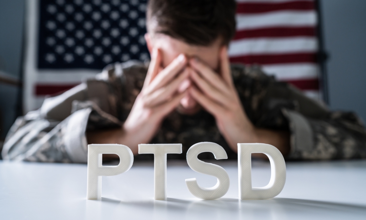 a soldier seated in front of an American flag with his head in his hands and the letters "PTSD" in front of him
