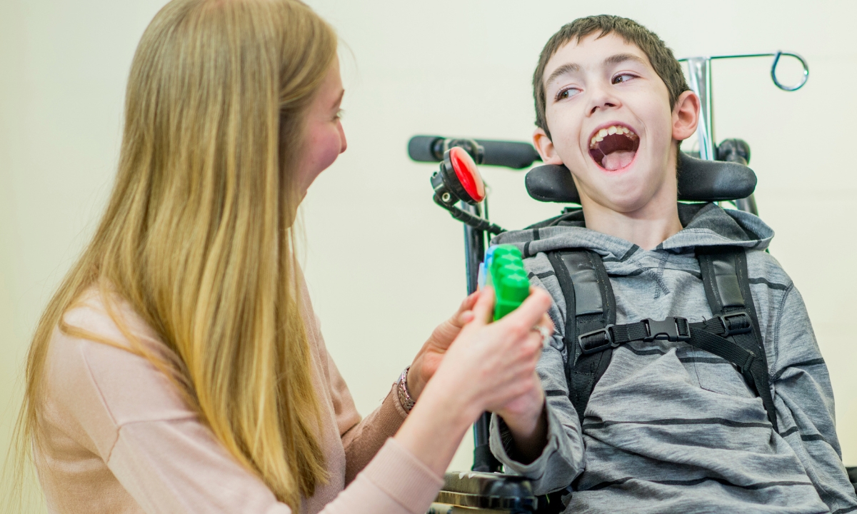 a young boy in a wheelchair smiling at his caregiver as she hands him a block
