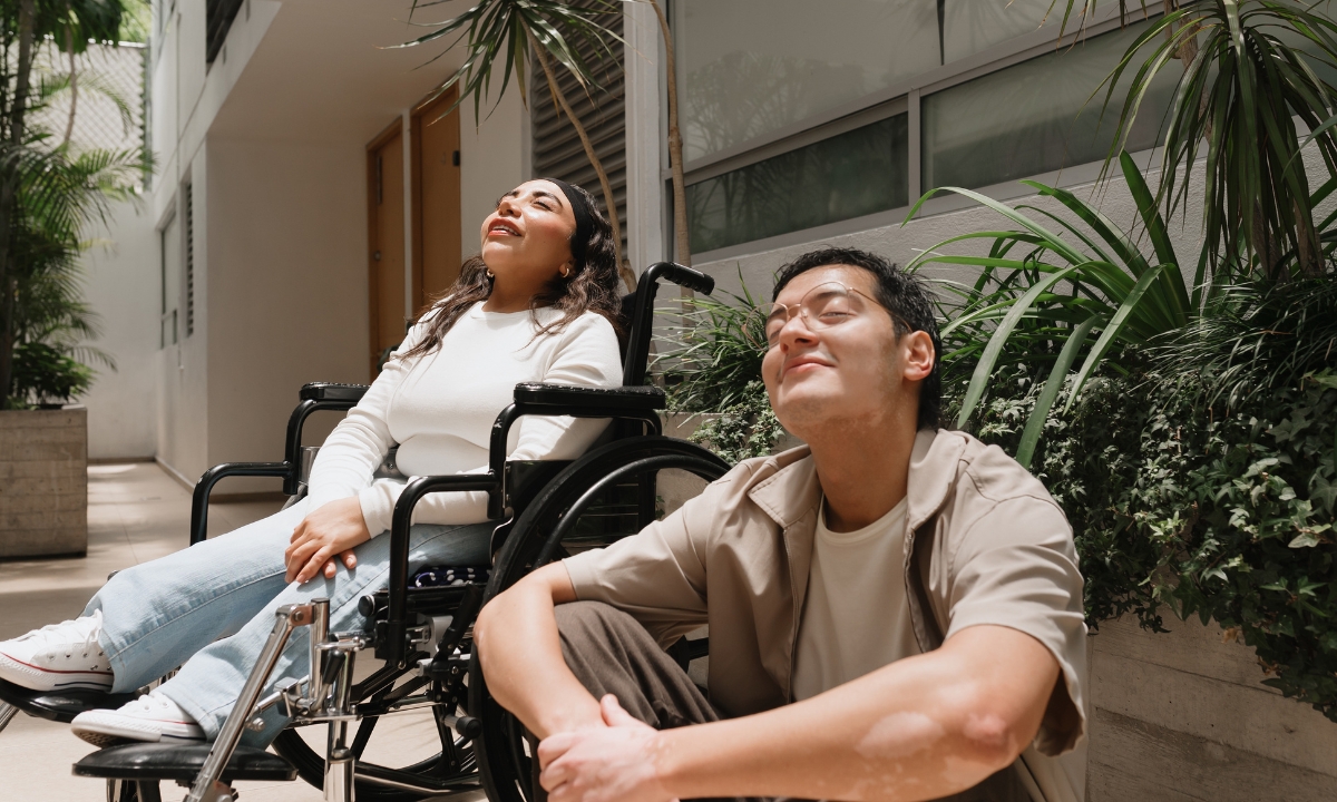 a man sitting next to a wheelchair-bound woman and they both are smiling and facing the sun with their eyes closed