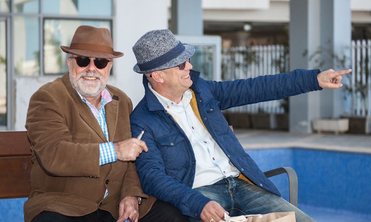 an elderly couple sitting next to each other on a bench, one lookig toward the camera and the other looking and pointing away