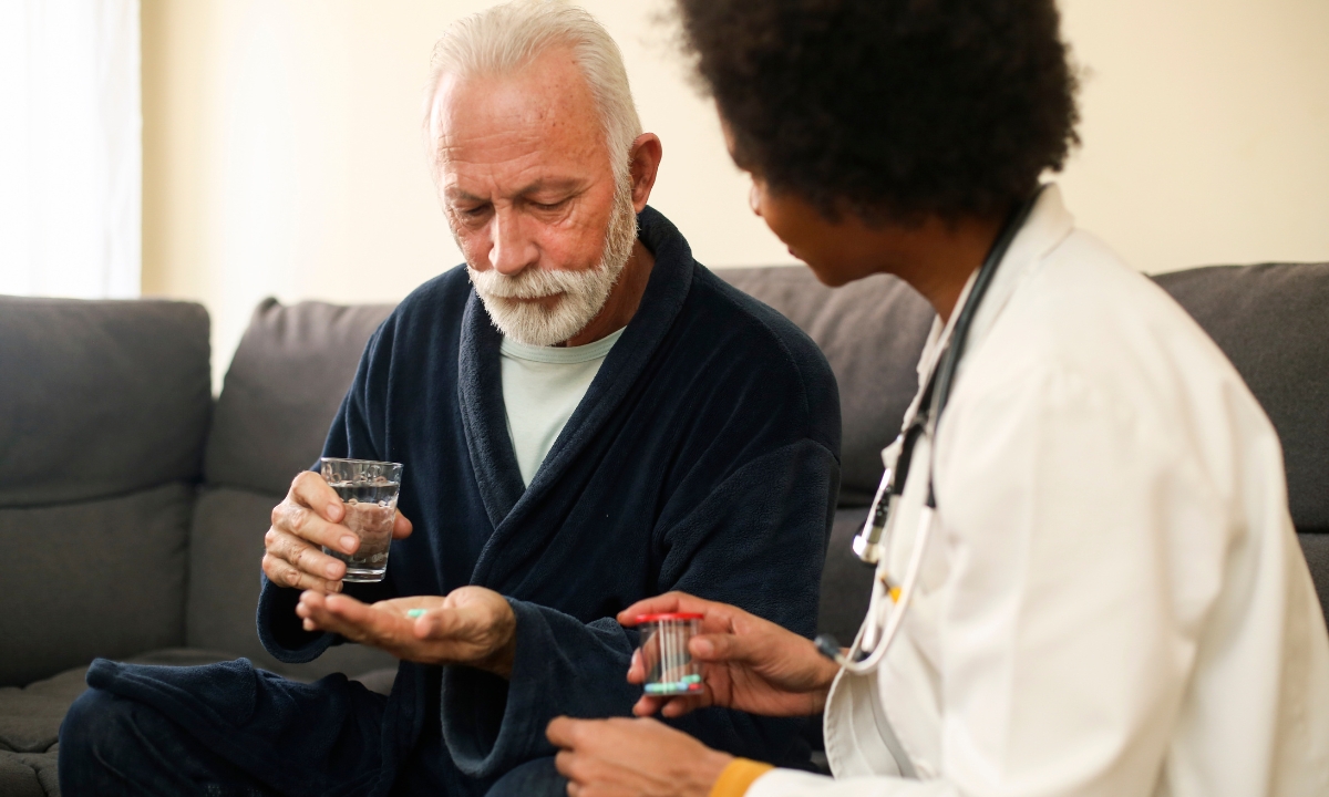 a doctor sitting on a couch with a senior patient and handing him medication