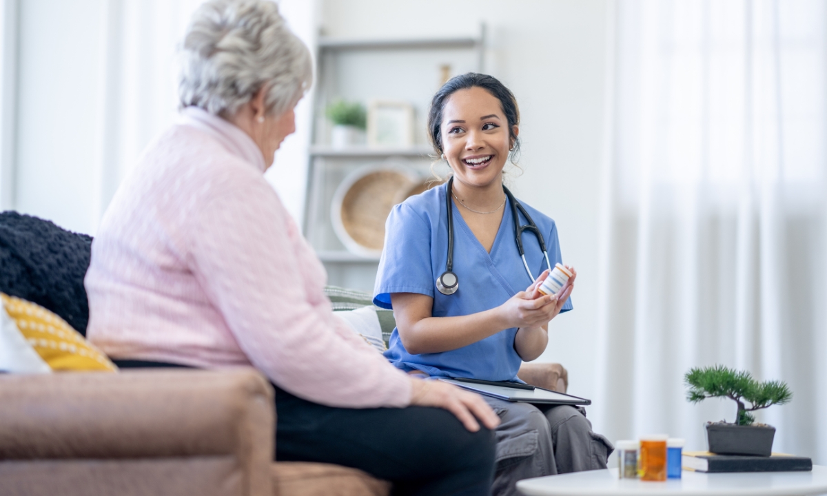 a nurse and an elderly patient sitting on a couch, the nurse shows the patient a bottle of prescription pills, both are smiling