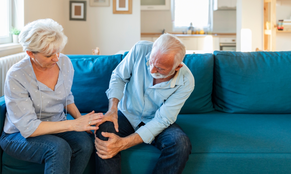 an elderly couple  sitting on a couch, one holding his knee and his wife holding him, too