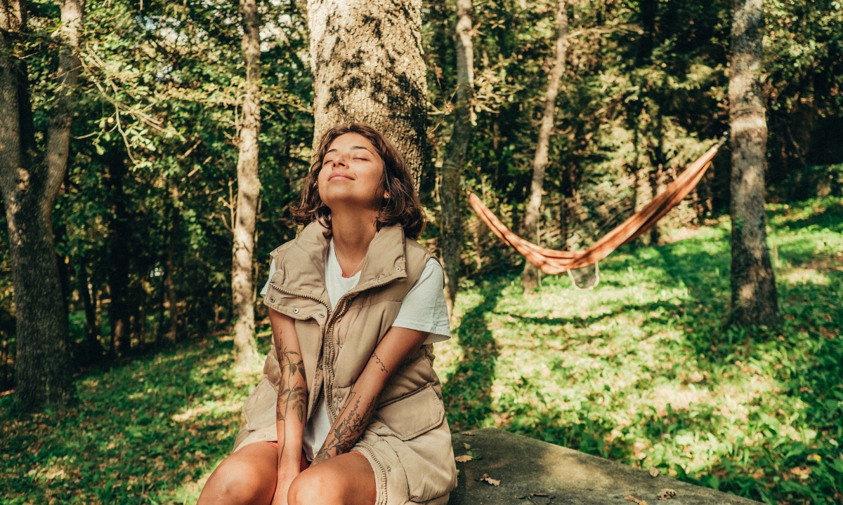 a woman in the forest, sitting at the base of a tree with her eyes closed and her face pointed up to the sun