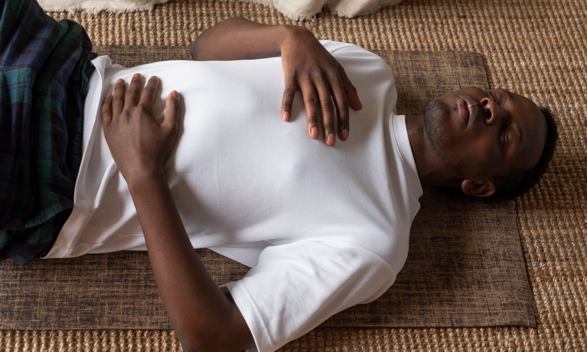 a man lying on his back on a mat on the floor, his eyes shut and his hands on his chest and stomach