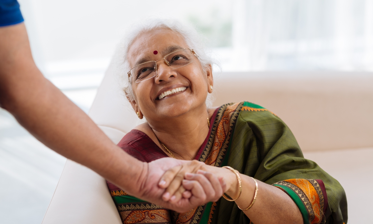 an elderly lady sitting on her couch, smiling up at and holding the hand of someone out of frame