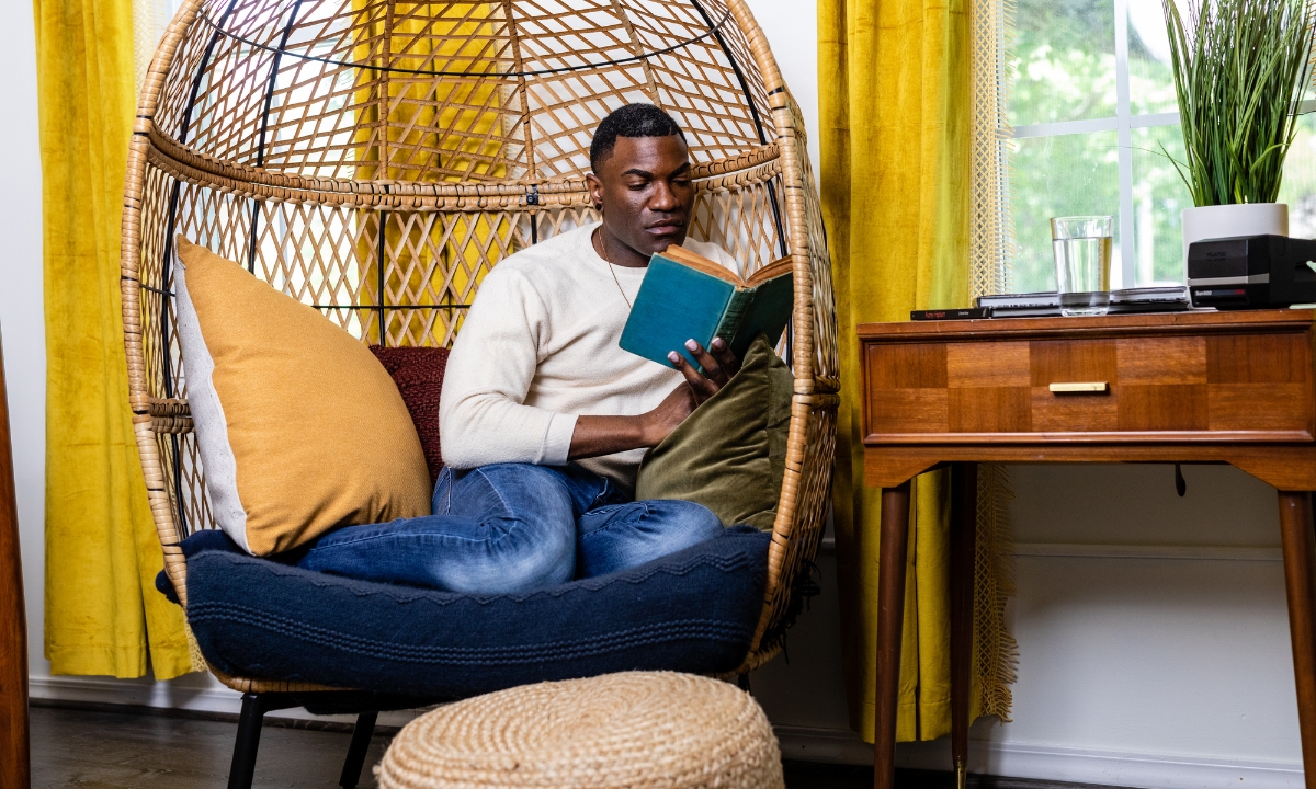 a man curled up in a wicker nest chair reading a book