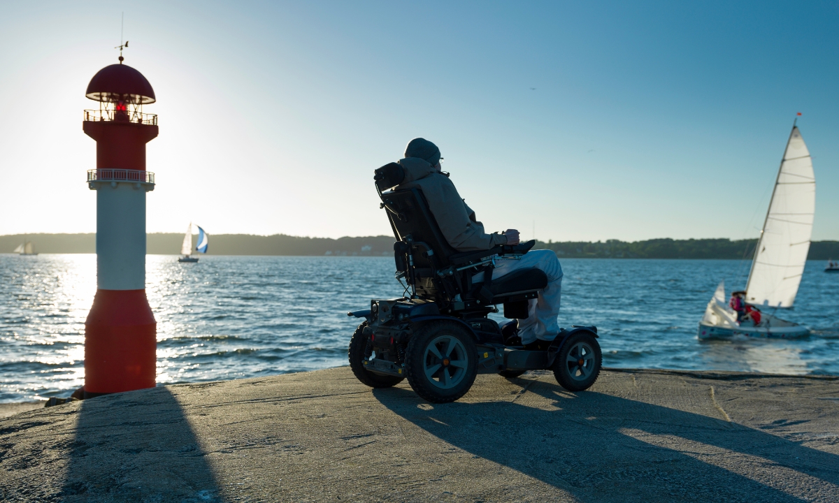 a person in a bulky coat in an electric wheelchair sitting by the ocean and looking out at the boats, sky, and tower buoy 