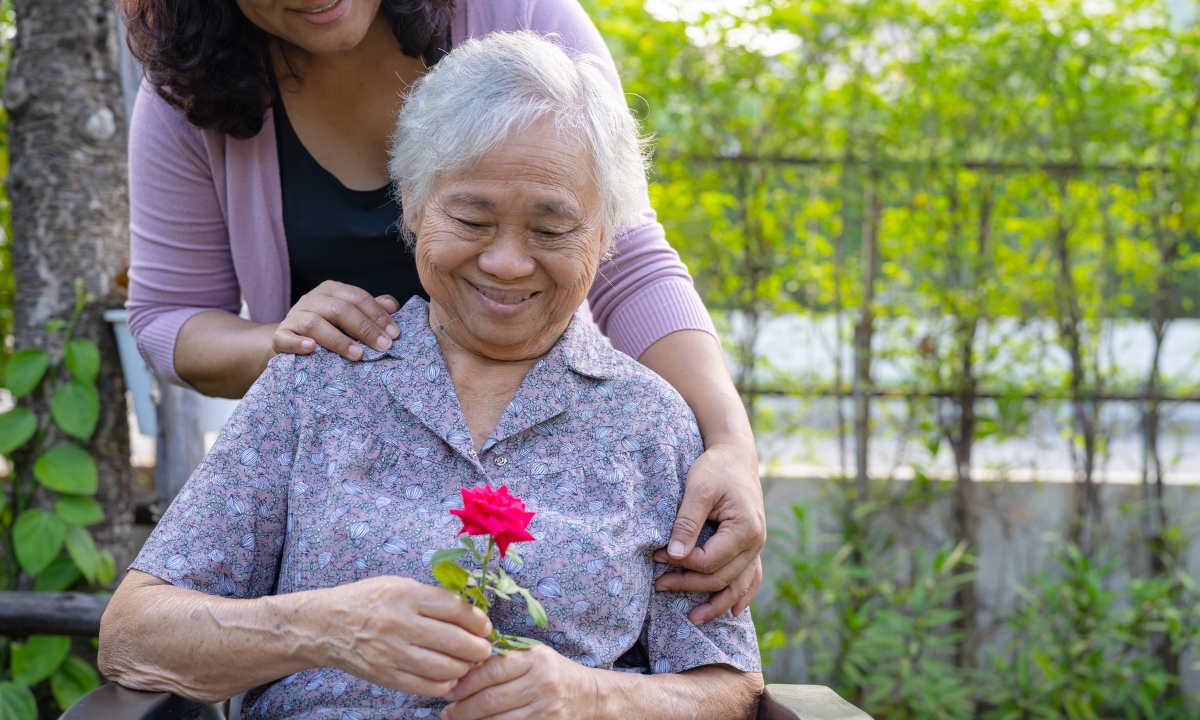 an elderly woman in a wheelchair holding a rose and smiling down at it; you can see another person behind her, their hands on her shoulders