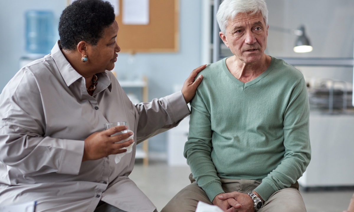 a woman next to a man, her hand on his shoulder as he looks at someone off camera and talks about something. he looks distressed and she looks concerned and supportive.