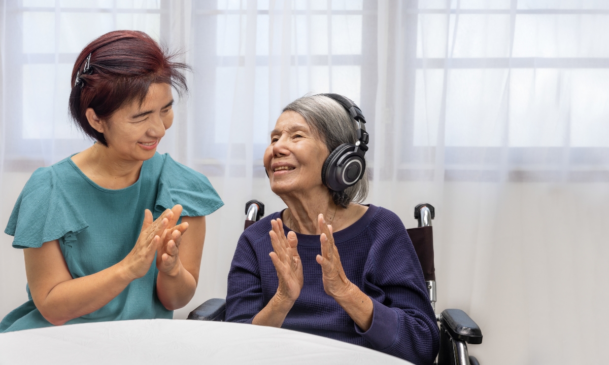 an in-home nurse clapping and dancing with an elderly woman who is wearing headphones; both are smiling widely.